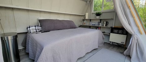 Ein Einzelbett mit gesteppter Bettdecke, ein Nachttisch mit Lampe, ein Mülleimer und ein Fenster mit Vorhängen.