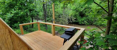 Une terrasse en bois avec un barbecue et un espace salon donnant sur une zone boisée traversée par un ruisseau.