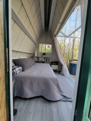A cozy attic bedroom with a bed, a small table, and a window overlooking trees.