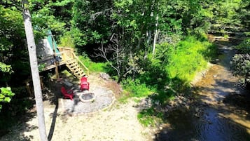A forested area with a wooden deck, a fire pit, and a small stream.