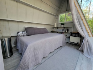 A single bed with a quilted cover, a bedside table with a lamp, a trash bin, and a window with curtains.