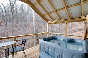 A hot tub on a wooden deck with a metal table and chair, surrounded by a metal railing, overlooking a forest.