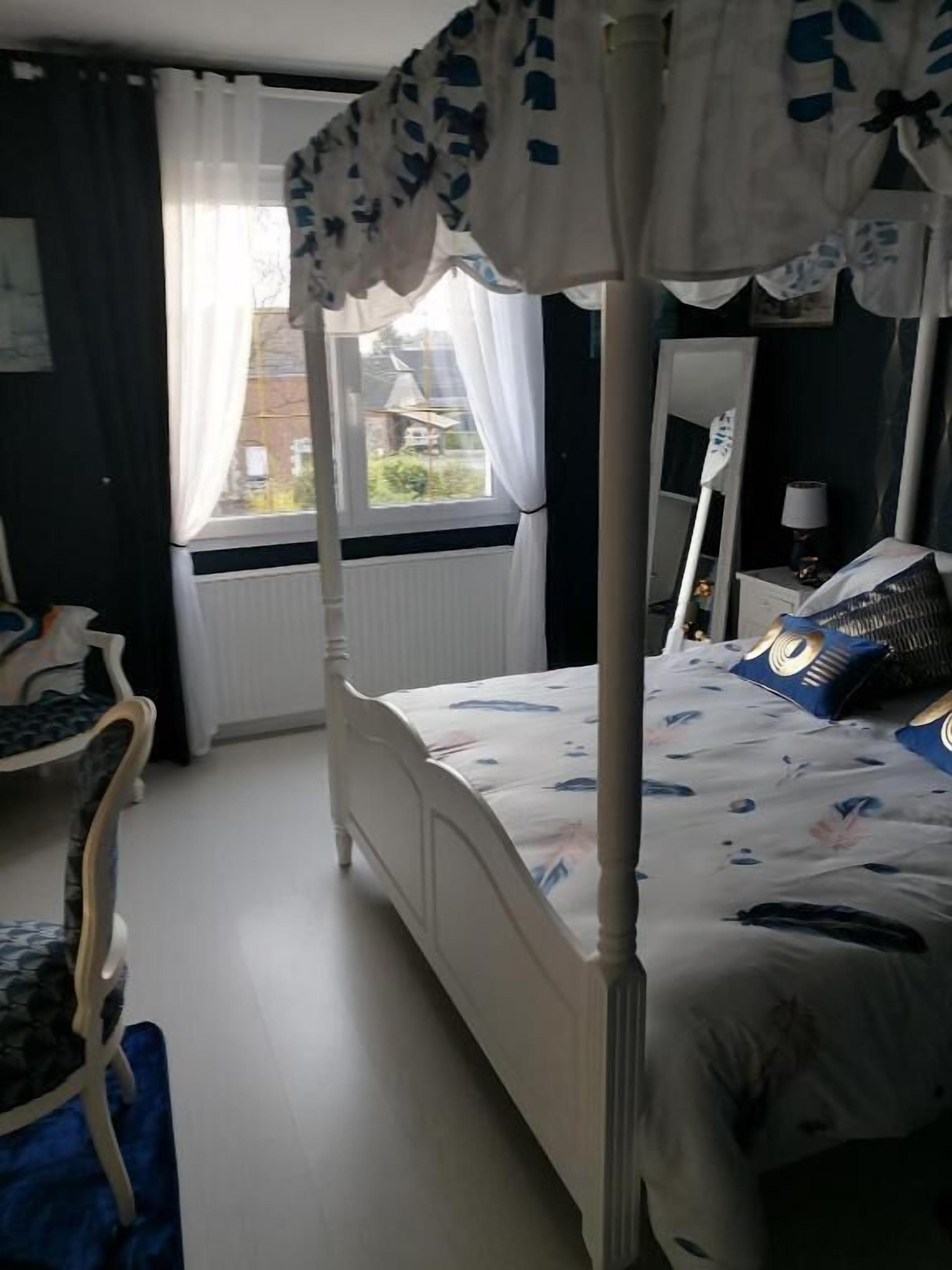 A bedroom with a four-poster bed, a window with curtains, a chair, a bedside table with a lamp, and a mirror.