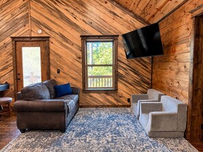Ein gemütliches Zimmer mit Holzbalkendecke, einem großen Fenster, einem Flachbildfernseher, einem braunen Sofa, einem grauen Sofa und einem gemusterten Teppich.