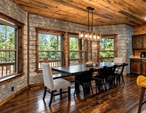 A dining area with a large table and chairs, wooden walls, and large windows. - Luxury Lodge with Hot Tub, Pool & Game Room near Broken Bow Lake – Hochatown, OK (Broken Bow)