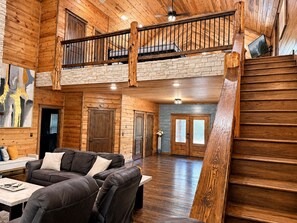 A spacious living area with a large sofa, a dining table, and a staircase leading to an upper level. - Luxury Lodge with Hot Tub, Pool & Game Room near Broken Bow Lake – Hochatown, OK (Broken Bow)