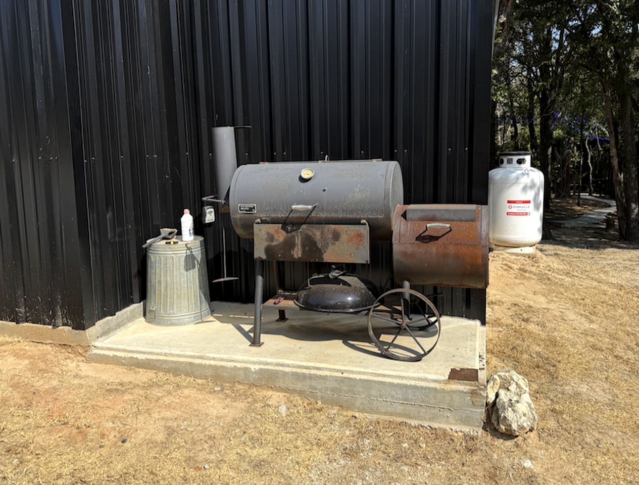 A portable grill with a gas tank and a metal container, placed on a concrete platform in front of a black metal building.