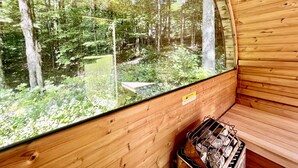 A wooden cabin with a large window offering a view of a forest. Inside, there is a bench and a fire pit. - Secluded Cottage with Stunning Sights in Winhall, Vermont (Winhall)