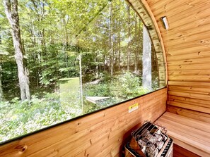 A wooden cabin with a large window offering a view of a forest. Inside, there is a bench and a fire pit. - Secluded Cottage with Stunning Sights in Winhall, Vermont (Winhall)