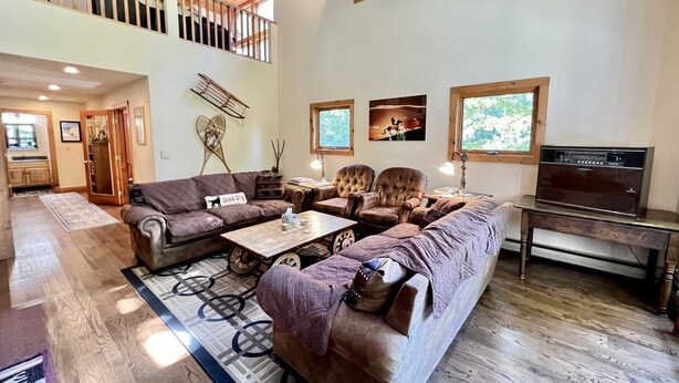 A spacious living room with a large sectional sofa, a coffee table, and a television. There is a balcony with wooden railings and a view of trees. - Secluded Cottage with Stunning Sights in Winhall, Vermont (Winhall)