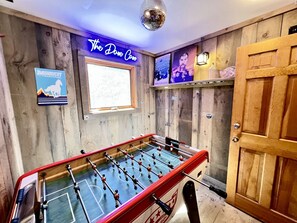Quarto com mesa de pebolim, paredes de madeira, uma luminária neon e uma bola de espelhos.