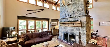 A spacious living room with a stone fireplace, wooden beams, and large windows.