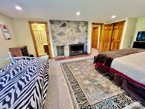 A bedroom with a stone fireplace, a bed with a patterned bedspread, a large rug, and wooden doors. - Secluded Cottage with Stunning Sights in Winhall, Vermont (Winhall)
