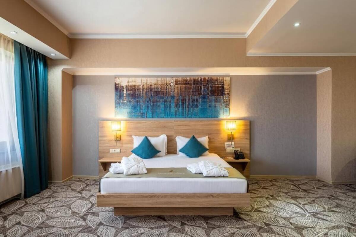 A hotel room with a large bed, two bedside lamps, a headboard with a cityscape artwork, and a patterned carpet.