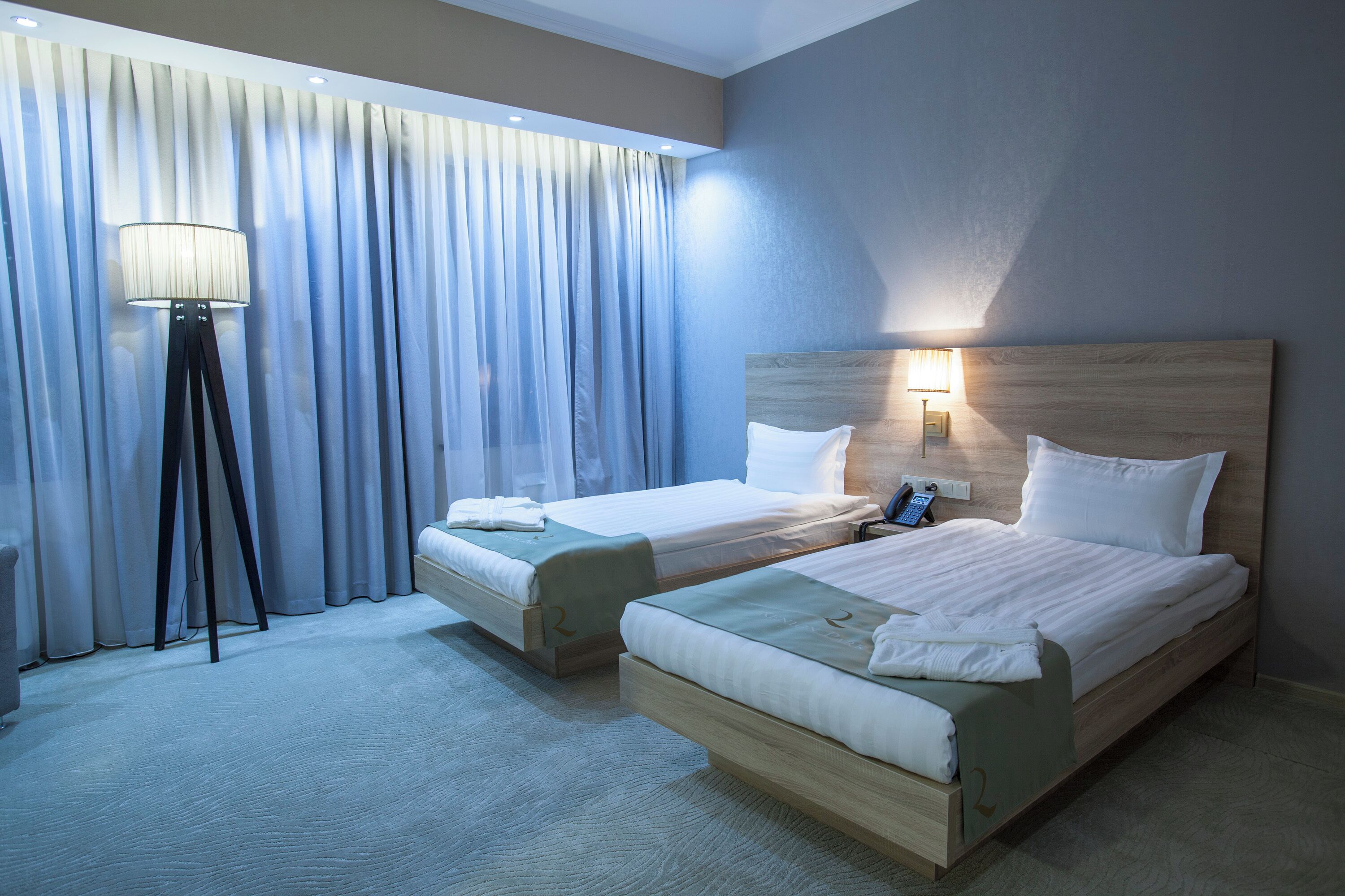 A hotel room with two beds, a floor lamp, a bedside table with a lamp, and a window with curtains.