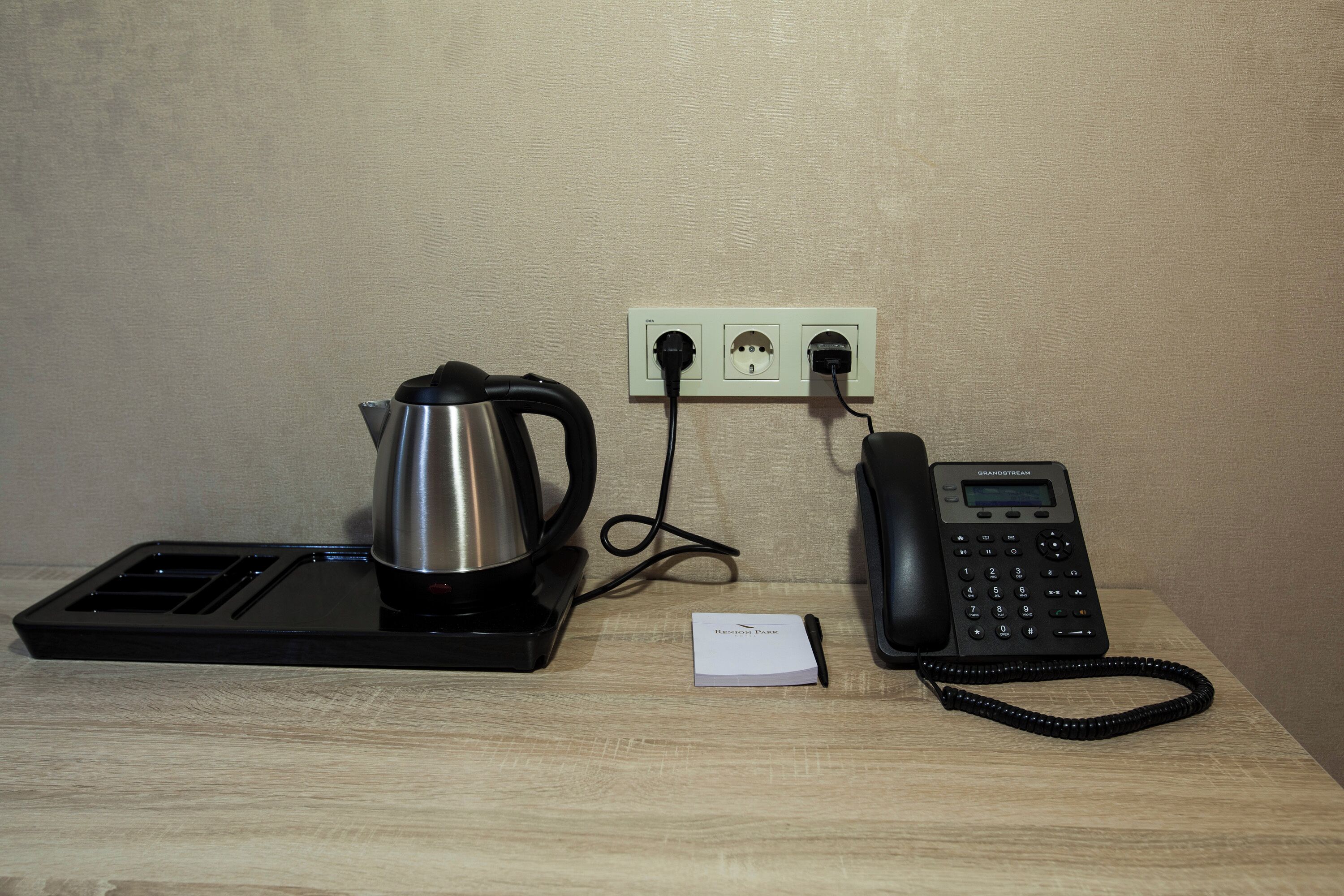 A kettle, a phone, and a power outlet on a wooden surface.