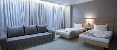 Una habitación de hotel moderna con sofá, dos camas individuales, una mesita de noche con lámpara y un ventanal con cortinas.