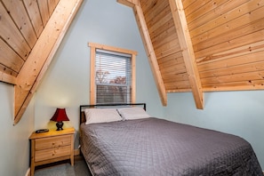 A bedroom with a sloped ceiling, a bed, a nightstand, a lamp, and a window with blinds.