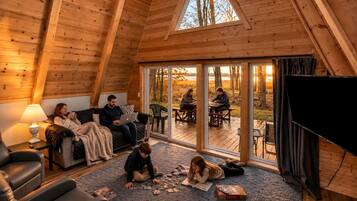 A cozy cabin interior with a wooden ceiling, a sofa, a dining table, and a large window overlooking a deck.