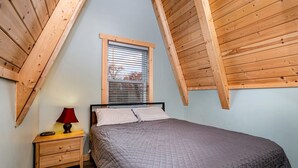 A bedroom with a sloped ceiling, a bed, a nightstand, a lamp, and a window with blinds. - Lakefront A-Frame with Lofted Interior Ideal for Outdoor Activities in Michigan (Au Gres)