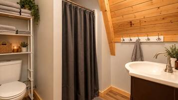 Ein Badezimmer mit Toilette, Waschbecken, einer Duschkabine, Regalen mit Handtüchern und einer Holzbalkendecke.