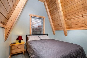 A bedroom with a sloped ceiling, a bed, a nightstand, a lamp, and a window with blinds.