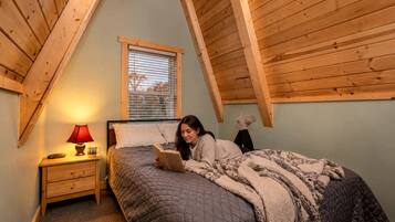 Un dormitorio con techo inclinado, una cama con colcha acolchada, una mesita de noche con lámpara y una ventana con persianas.