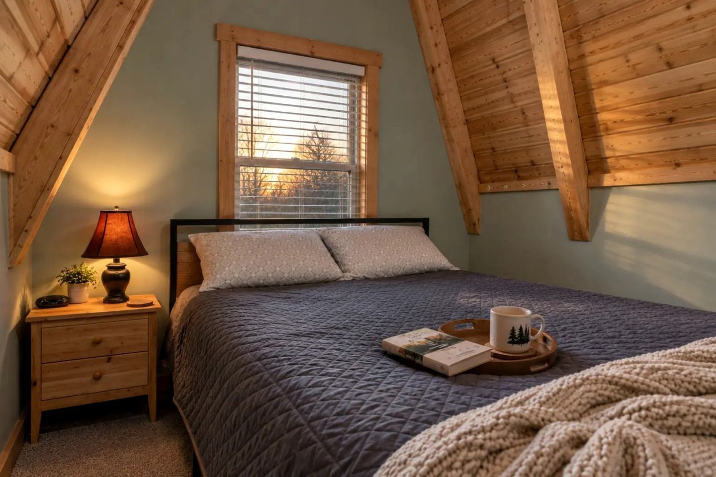 Un dormitorio con cama, mesita de noche con lámpara, ventana y vista a árboles fuera.