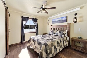 A bedroom with a bed, a ceiling fan, a window with curtains, a nightstand, and a wall-mounted lamp. - Stunning Old West Cabin with Fireplace in Island Park, Idaho (Island Park)