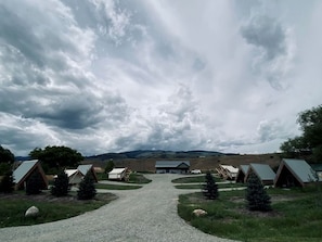 Un terrain de camping comprenant plusieurs tentes et cabanes, un sentier de gravier menant à un bâtiment central, et un paysage montagneux en arrière-plan.