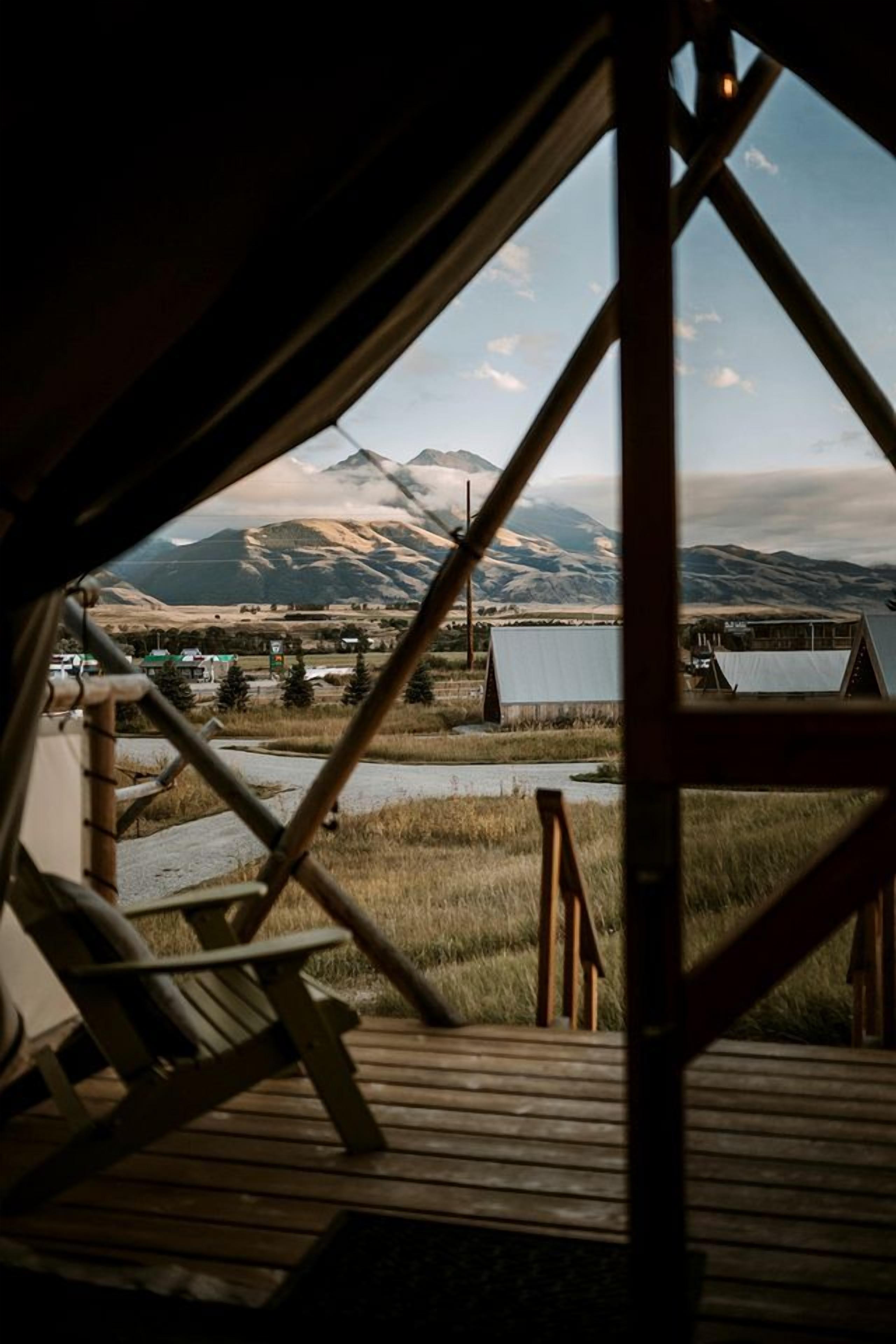 A view from inside a cabin with a wooden deck, a chair, and a mountain landscape in the background.