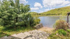 A lakeside view with a stone pathway leading to a dock, surrounded by greenery and hills.