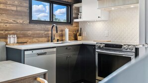 A modern kitchen with wooden wall paneling, a window, a sink, a dishwasher, a stove, and white cabinets. - Mont Orignal | Private Hot Tub | Alpenglow (Lac-Etchemin)