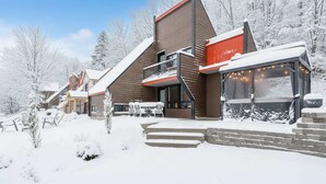 A modern lodge with a prominent red and brown exterior, a balcony, and a large window with string lights. - Mont Orignal | Private Hot Tub | Alpenglow (Lac-Etchemin)