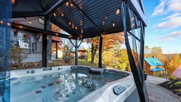 A hot tub with a glass enclosure, a covered deck, and a view of a house and trees. - Mont Orignal | Private Hot Tub | Alpenglow (Lac-Etchemin)