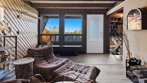 A cozy room with a wooden ceiling, a large window with a view of autumn trees, a bench, and a staircase leading to another level. - Mont Orignal | Private Hot Tub | Alpenglow (Lac-Etchemin)