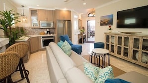 A modern kitchen with stainless steel appliances, a dining area with a white sofa and blue chairs, and a flat-screen TV mounted on the wall. - The Rod and Reel Resort-Grand Suite 6 (Anna Maria)