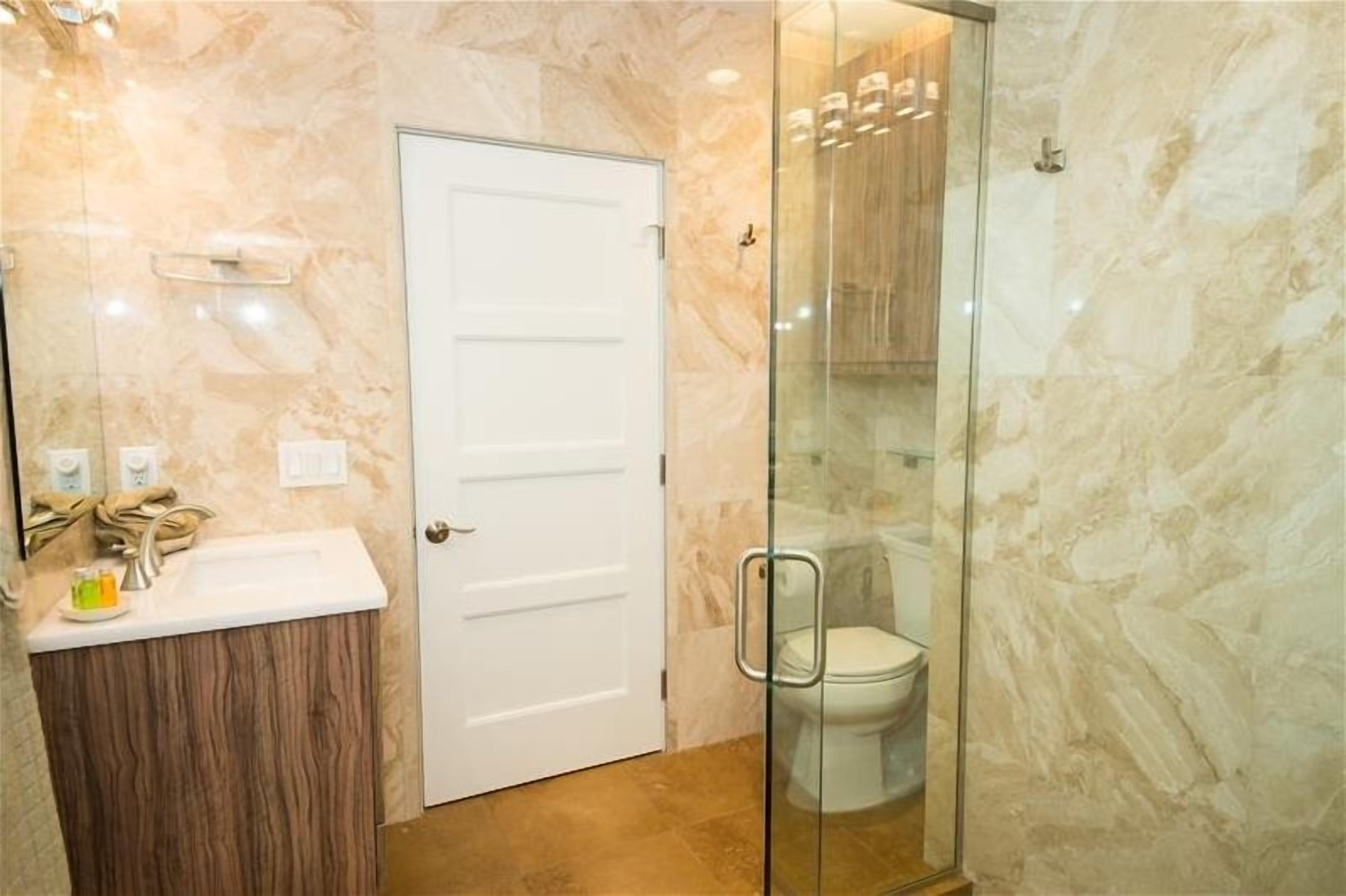 A bathroom with a marble wall, a white door, a glass shower enclosure, and a wooden vanity.