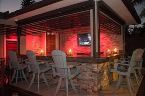 An outdoor bar area with Adirondack chairs, a stone wall, and a TV mounted on the wall. - The Rod and Reel Resort-Grand Suite 6 (Anna Maria)