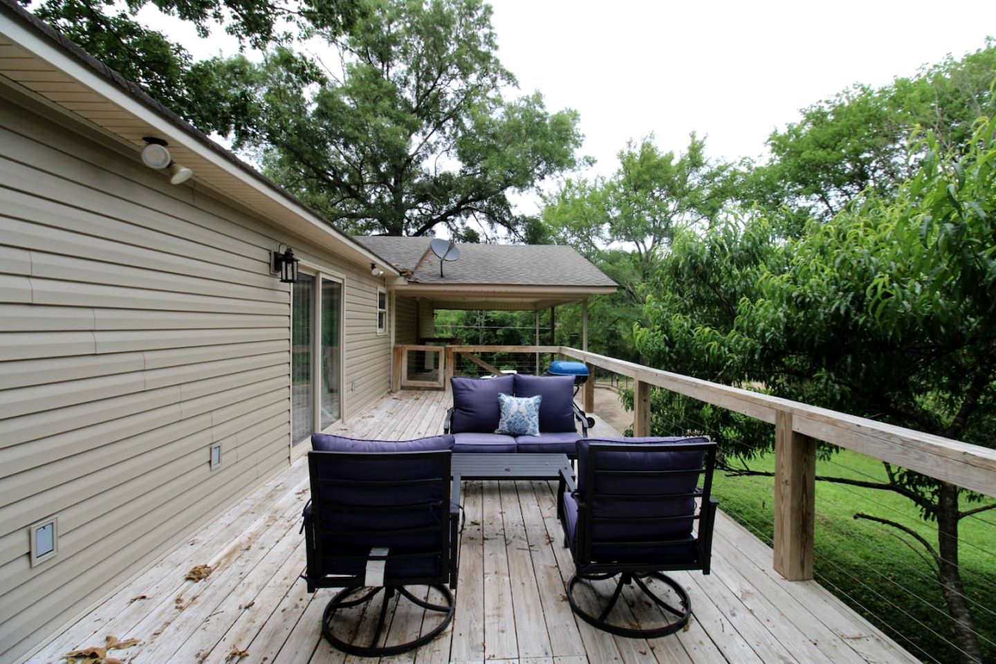 A covered outdoor deck with a sofa and chairs, a barbecue grill, and a wooden railing.