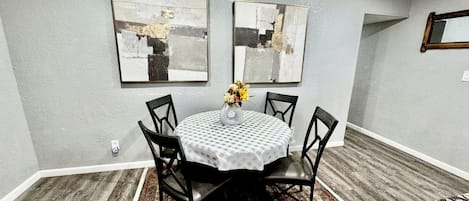 A dining area with a round table, black chairs, a white tablecloth, and two abstract paintings on the wall.