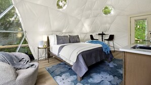 A tented accommodation with a bed, bedside table, lamp, chairs, and a small kitchen area.
