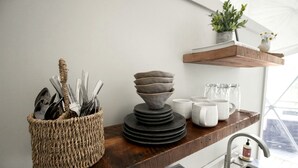 A kitchen counter with a woven basket containing utensils, stacked bowls, and white mugs on a wooden shelf.