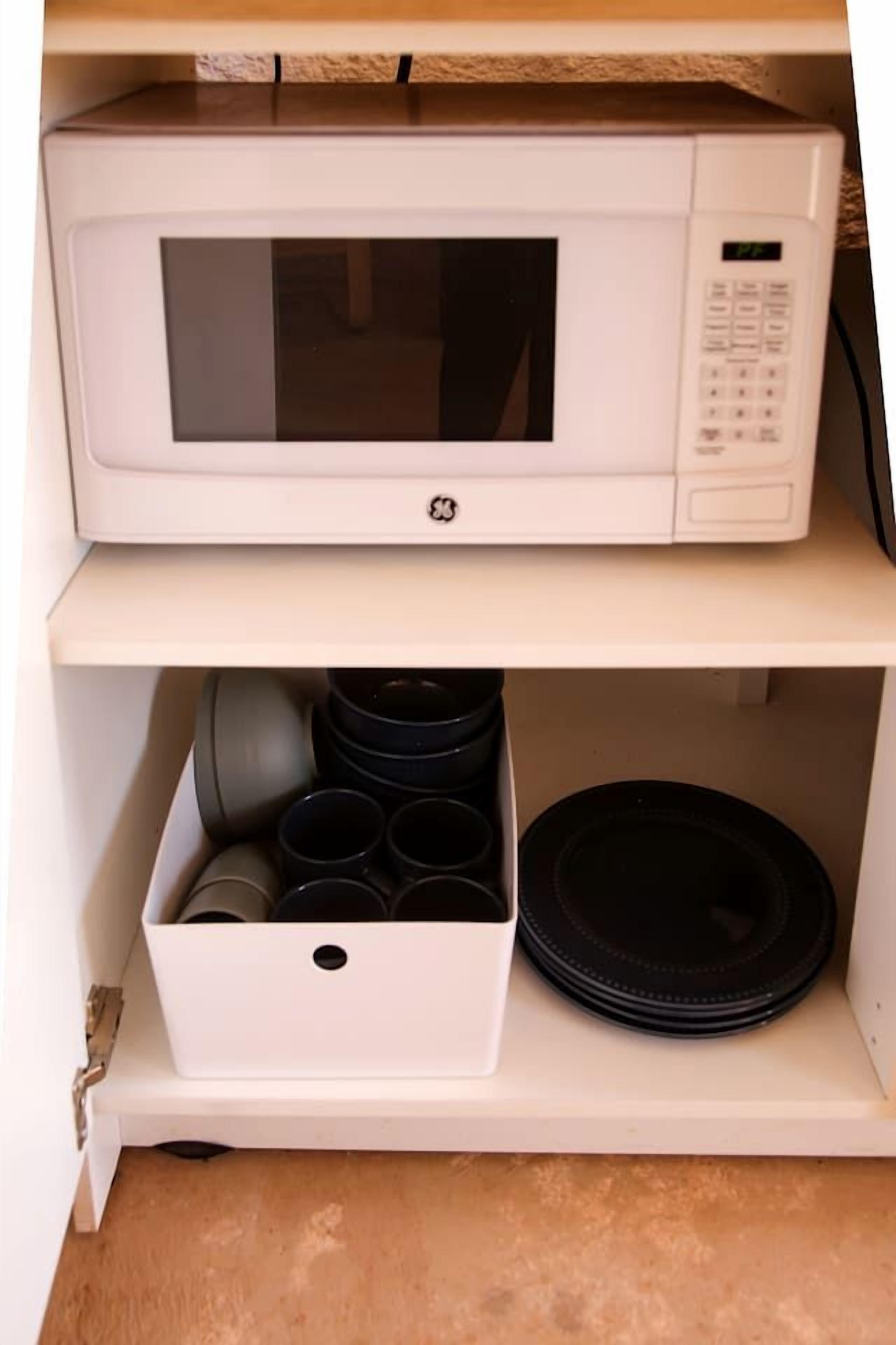 A white microwave on a shelf above a storage box containing black cups and plates.