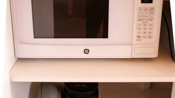 A white microwave on a shelf above a storage box containing black cups and plates.