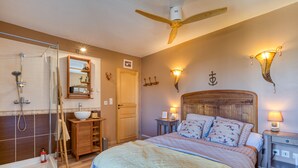 A bedroom with a wooden bed, a ceiling fan, a bedside table with a lamp, a nightstand, a wooden wardrobe, a mirror, and a shower area. - Guest Room "La Petite Camargue" with Private Pool, Shared Terrace and Wi-Fi (Vauvert)