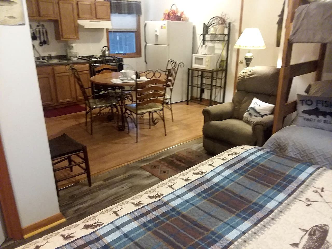 Une pièce de taille modeste comprenant un lit, une table à manger avec des chaises, un coin cuisine avec des placards et des appareils électroménagers, et un petit espace salon avec un fauteuil.