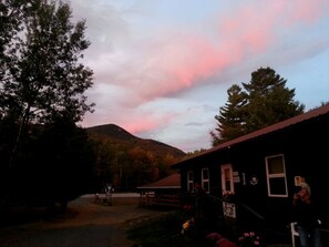 A rustic building with a dirt path leading to it, surrounded by trees and a mountain in the background.