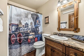 A bathroom with a vintage train-themed shower curtain, a white toilet, a sink with a marble countertop, and a wooden vanity. - Delightful Old West Cabin in Island Park, Idaho (Island Park)
