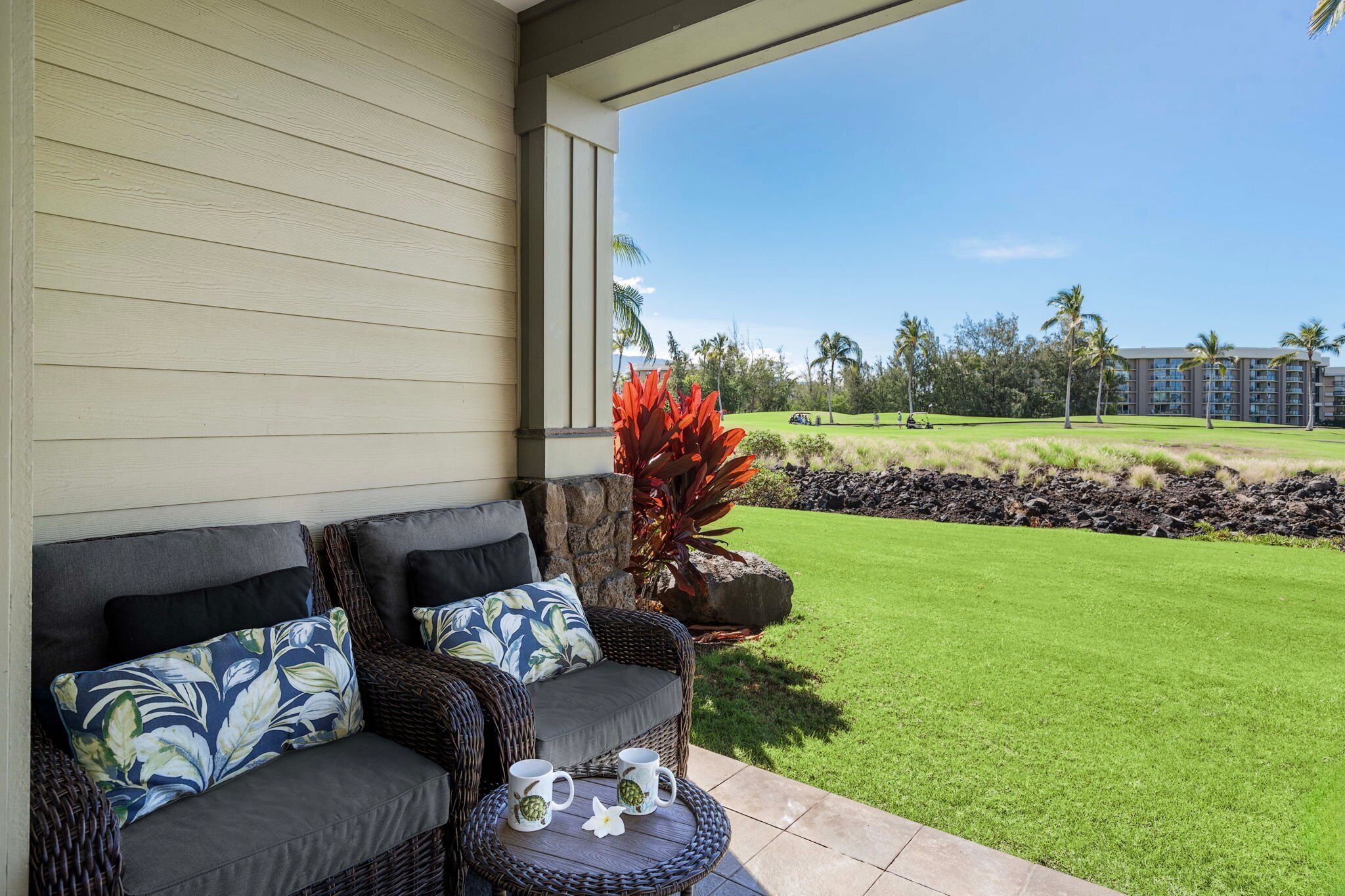 A patio with wicker furniture, a stone planter with red foliage, and a view of a golf course and hotel in the background.